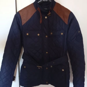 Quiltad jacka från Ralph Lauren - Säljer en snygg quiltad jacka från Ralph Lauren i mörkblått med bruna detaljer på axlarna. Jackan har en stilren design med knappar och ett bälte i midjan för en perfekt passform. Den är tillverkad i polyester och passar perfekt för höst och vår. Perfekt för dig som vill ha en klassisk och tidlös stil!