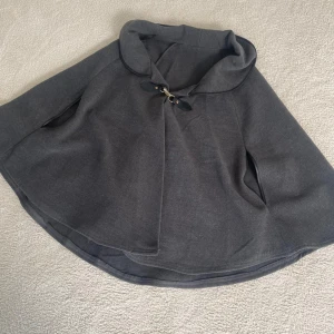 Grå cape med knäppning - Säljer en elegant grå cape med en snygg knäppning framtill. Den har en mjuk och bekväm passform, perfekt för höst och vår. Capen är i ett stilrent och tidlöst snitt som passar till många olika outfits.