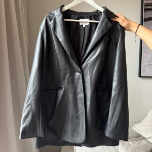 Svart skinn-liknande kavaj - Snygg svart skinnkavaj i mycket bra skick. Den har en klassisk design med en knapp framtill och långa ärmar. Perfekt för höst och vår, och funkar både till vardags och fest. Storleken är normal och materialet är mjukt och bekvämt. Nypris 700 kr