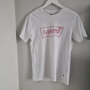 Vit t-shirt från Levi's - Säljer en klassisk vit t-shirt från Levi's i bomull. Den har en röd logga på framsidan och i bra skick (förutom knappt synliga fläckar, se bild) Perfekt för en avslappnad stil eller som basplagg i garderoben. Kortärmad och normal passform.