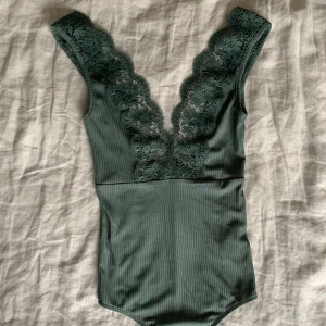 Grön ribbad bodysuit med spetsdetaljer - Snygg grön bodysuit i ribbat material med vackra spetsdetaljer längs axelbanden och ner över bröstet. Perfekt för en kväll ute eller en speciell tillställning. Den har en tight passform som framhäver figuren.
