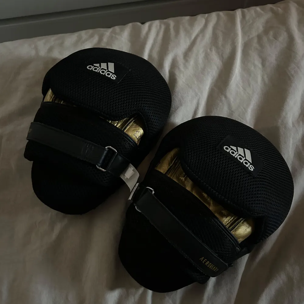 Säljer ett par svarta Adidas boxning mittsar i nytt skick. De har en snygg design med guldiga detaljer och justerbara remmar för perfekt passform. . Övrigt.