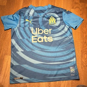 Blå fotbollströja Marseille (OM) - Säljer en snygg blå fotbollströja från Puma med korta ärmar. Tröjan har olika blå nyanser och loggor på bröstet. Perfekt för fotbollsfans eller träning! 🏆
