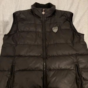 Svart dunväst från EA7 - Säljer en snygg svart dunväst från EA7 i mycket bra skick. Västen har en dragkedja framtill och två praktiska fickor med dragkedjor. Perfekt för höst och vår när du vill hålla dig varm men ändå se stilren ut. Passar både till vardags och lite finare tillfällen.