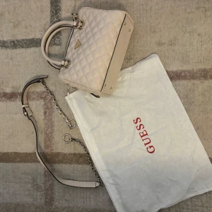 Beige quiltad handväska från Guess - Säljer en superfin beige handväska från Guess i mycket bra skick. Väskan är quiltad med snygga detaljer i silver och har både handtag och en avtagbar axelrem. Perfekt för både vardag och fest! 👜✨