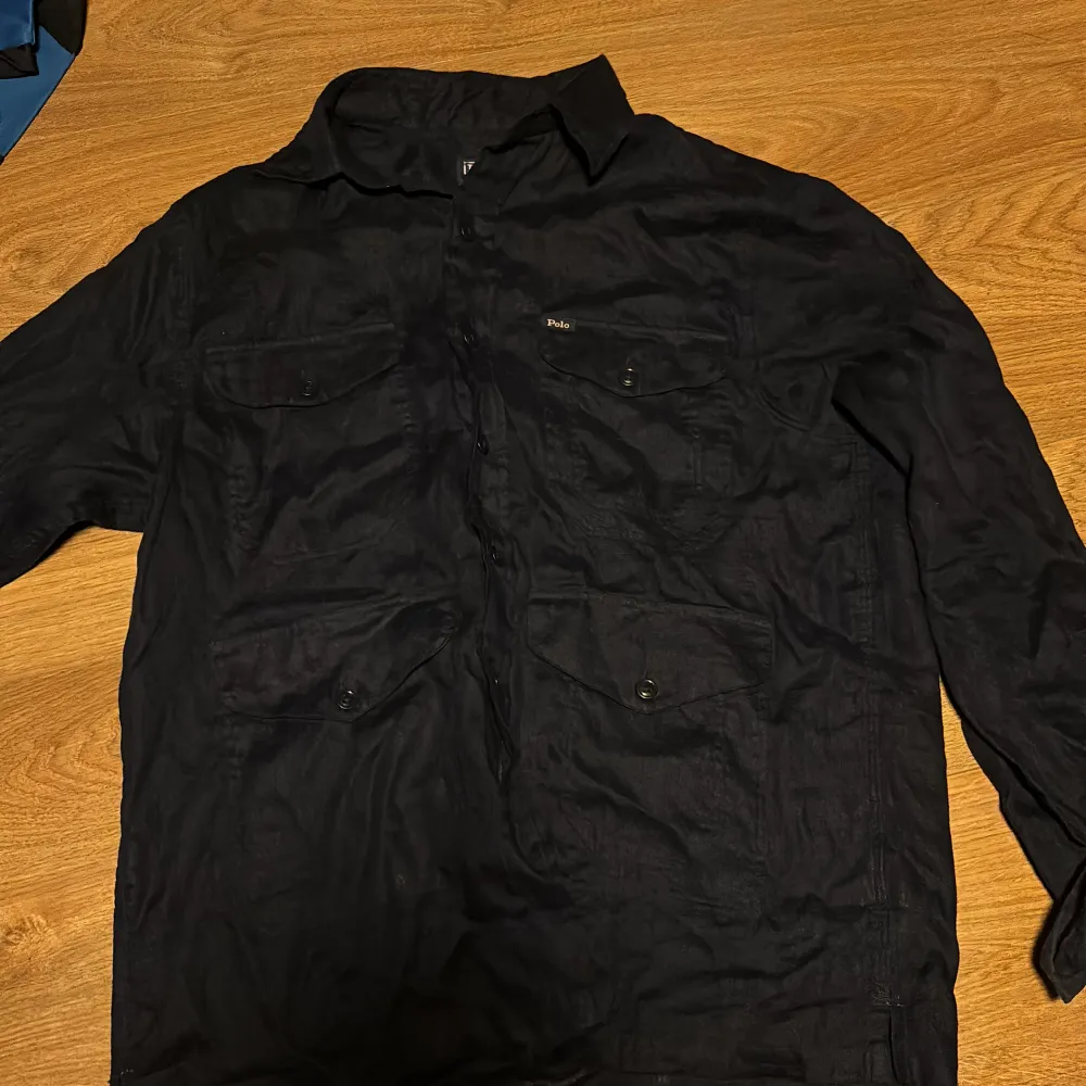 Säljer en snygg mörkblå overshirt från Polo Ralph Lauren i linne. Perfekt för höst och vår med sina långa ärmar och stilrena knappar. Skjortan har praktiska fickor framtill och en klassisk krage. Passar både till vardags och lite finare tillfällen. Qr koden funkar som man kan se på första bilden där det står authenticated. Den är i storlek s och i princip aldrig använd.. Skjortor.