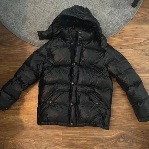 Svart dunjacka från Polo Ralph Lauren - Säljer en svart dunjacka från Polo Ralph Lauren i bra skick. Jackan har en broderad blå logga på bröstet och är långärmad med luva, skulle säga att den är lite kort i armarna. Dm mig vid mer frågor eller bilder😼. Armlängd 62cm, Längd jacka 67cm, Vidd bröst 110cm, Vidd midja 110cm