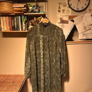 Grön mönstrad klänning från H&M - Säljer en grön klänning från H&M med ett ormskinnsmönster. Klänningen har långa ärmar och en hög krage, perfekt för höst och vinter. Materialet känns mjukt och bekvämt, och den har en lös passform som gör den skön att bära hela dagen.