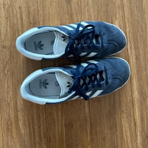 Adidas Gazelle i blå mocka - Säljer ett par snygga Adidas Gazelle i blå mocka med vita detaljer. Skorna har klassiska tre ränder på sidan och texten 'GAZELLE' i guld. De är i bra skick och perfekta till vardags. Skostorlek dam 38.