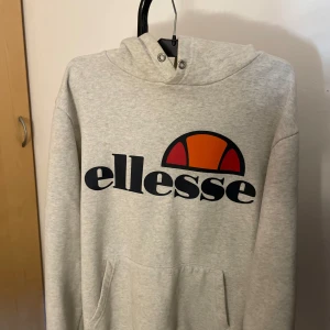 Grå hoodie från Ellesse - Säljer en snygg och bekväm grå hoodie från Ellesse. Den har en stor logga på framsidan i svart, rött och orange. Perfekt för en chill dag eller när du vill ha en sportig look. Den har en klassisk känguruficka och en skön huva. Men sakanr tyvär snöret som skall gå igenom luvan