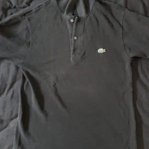 Svart pikétröja från Lacoste - Säljer en klassisk svart pikétröja från Lacoste med den ikoniska krokodilen broderad på bröstet. Tröjan har en knappslå med två knappar och är i mycket bra skick. Tröjan säljes för endast 170kr! 🐊