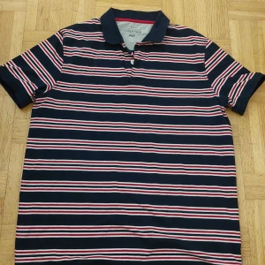 Randig pikétröja från Essentials - Säljer en snygg randig pikétröja från Essentials i storlek XL. Tröjan har en klassisk krage och knappar framtill. Färgerna är marinblå, vit och röd, vilket ger en stilren look. Perfekt för både vardag och lite mer uppklädda tillfällen. Passar bra till jeans eller chinos!