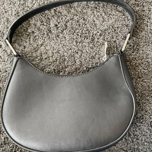 Svart axelväska i skinn - Säljer en stilren svart axelväska i skinn med dragkedja. Väskan har en enkel och elegant design, perfekt för både vardag och fest. Den är i mycket bra skick. FEJK SKINN