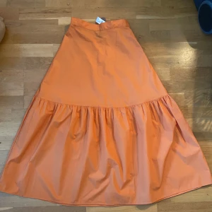 Orange/aprikos kjol från Lindex - Säljer en superfin orange kjol från Lindex i storlek 34. Kjolen har en härlig volangdetalj och är perfekt för sommardagar. Den är gjord i ett lätt och bekvämt material, vilket gör den idealisk för både vardag och fest. 