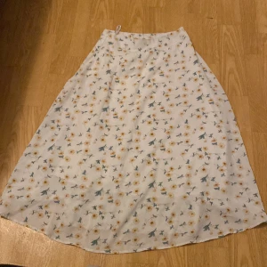 Vit blommig kjol - Säljer en superfin vit kjol med blommigt mönster i olika färger. Perfekt för vår och sommar! Kjolen är i ett lätt och luftigt material, vilket gör den idealisk för varma dagar. Den har en normal passform och är i mycket bra skick. Passar perfekt till både vardag och festliga tillfällen.