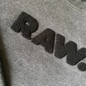 Grå sweatshirt från G-Star RAW - Säljer en snygg grå sweatshirt från G-Star RAW i storlek S. Tröjan har en cool svart text 'RAW.' på bröstet och är i mycket bra skick. Perfekt för en avslappnad stil och passar till alla årstider. Skön och stilren design!