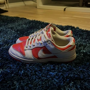 Nike Dunk i rött och vitt - Säljer ett par Nike Dunk i rött och vitt. Skorna har en klassisk design med snörning och är i bra skick. Perfekta för både vardag och träning. Sulan är röd och ovandelen har en snygg kombination av rött och vitt. Lät ai skriva och det stämmer väldigt bra. Dom är i väldigt bra skick och säljs pga pengar behov. Köptes för 200 av min polare och det är därför jag säljer billigt, Såklart är dom äkta så tveka inte på att höra av er