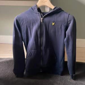 Säljer en snygg mörkblå hoodie från Lyle & Scott i juniorstorlek 10/11 år. Den har en dragkedja framtill och en gul logga på bröstet. Perfekt för höst och vår, och den är i bra sjikts. Passar både till skolan och fritiden! Ops den är lite blekt!