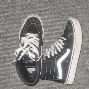 Svarta höga sneakers från Vans - Säljer ett par svarta höga sneakers från Vans i bra skick. De har vita snören och den klassiska vita sidostripen. Sulan är brun med ett rutmönster för bra grepp. Perfekta för vardagsbruk och streetwear-stil.