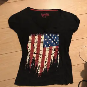 Säljer en svart t-shirt från Superdry med ett coolt amerikanskt flaggmotiv i rött, vitt och blått. T-shirten har en v-ringad hals och korta ärmar. Passar bra till jeans eller shorts!