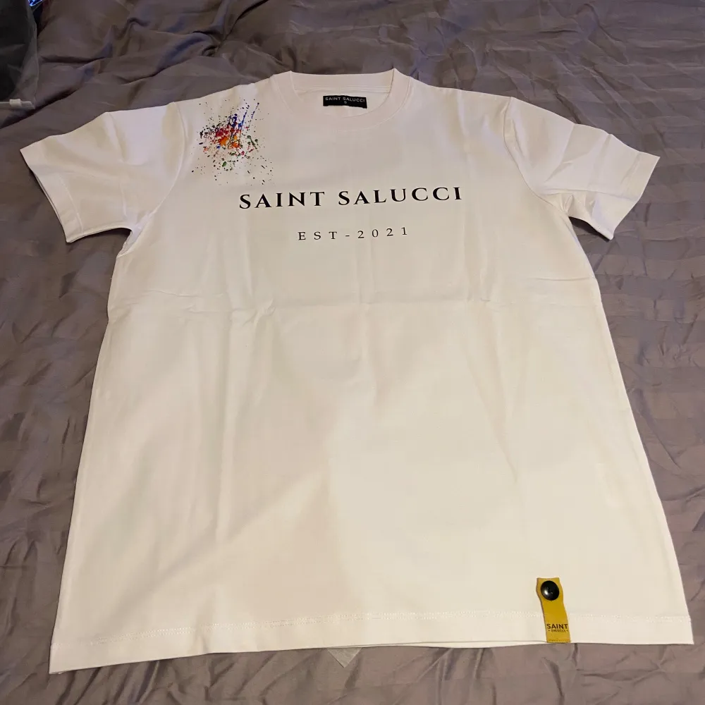 Säljer en snygg vit t-shirt från Saint Salucci i storlek S.M.L.XL Den har ett coolt färgstänk-mönster på axeln och texten 'SAINT SALUCCI EST. 2021' tryckt på framsidan. Perfekt för en avslappnad stil eller en utekväll. T-shirten är i nytt skick och gjord av mjukt bomullsmaterial.. T-shirts.