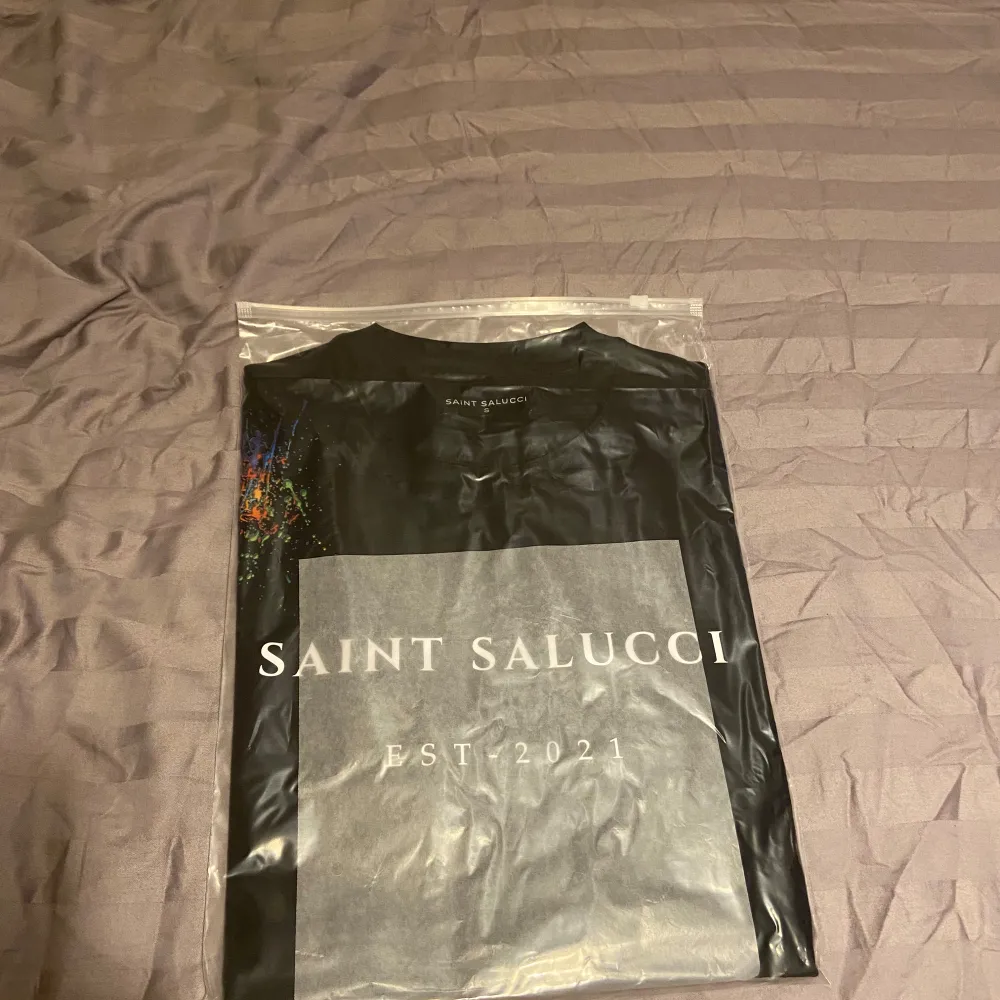 Snygg svart t-shirt från Saint Salucci med tryck 'EST. 2021' på framsidan. Den har en cool färgexplosion på axeln och en gul detalj med knapp längst ner. Perfekt för en casual look! Storlek S.M. L . XL . T-shirts.