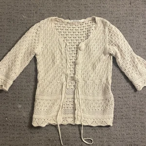 Beige stickad kofta från RoseBud - Säljer en superfin beige stickad kofta från RoseBud i storlek XS. Den har ett vackert hålmönster och knappar framtill. Koftan har trekvartsärmar och ett knytband i midjan för en söt look. 