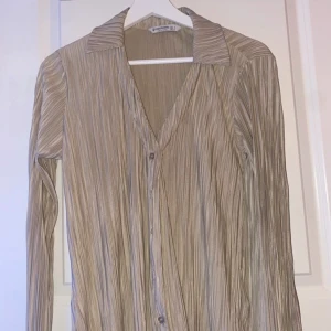 Beige långärmad blus - Säljer en elegant beige blus med långa ärmar och ett diskret randigt mönster. Blusen har en klassisk krage och knappar framtill. Perfekt för både vardag och festliga tillfällen. Passar bra till höst och vår.