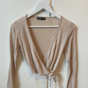 Beige omlottopp från New Yorker - Säljer en superfin beige omlottopp från New Yorker. Den har en v-ringad design och knytning i midjan som ger en snygg passform. Perfekt för både vardag och fest! 💫