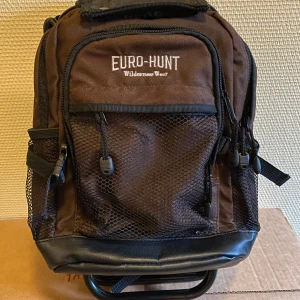 Brun ryggsäck med inbyggd stol från Euro-Hunt - Praktisk brun ryggsäck från Euro-Hunt med inbyggd stol, perfekt för utflykter och jakt. Ryggsäcken har flera fickor med dragkedja och nät för extra förvaring. Den är tillverkad i slitstarkt material och har justerbara remmar för bekväm passform.
