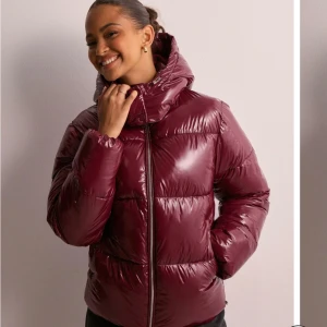 Vinröd pufferjacka - Säljer en snygg vinröd pufferjacka i mycket bra skick. Jackan har en hög krage och dragkedja framtill, perfekt för kyliga dagar. Den är långärmad och har en glansig finish som ger en trendig look. Perfekt för både höst och vinter!