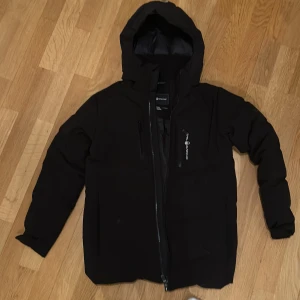 Svart dunjacka från Sail Racing - Säljer en svart dunjacka från Sail Racing i mycket bra skick. Jackan har en dragkedja framtill och en praktisk huva. Den är perfekt för kalla dagar med sitt 500 FP dunfoder. Passar både till vardags och utomhusaktiviteter. Storlek 160-170 cm den har dock ett hål vid fickan som dock är lagat knappt använd pris kan diskuteras