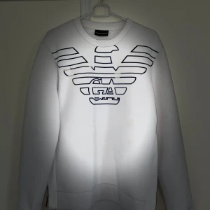 Vit tröja från Emporio Armani - Säljer en stilren vit tröja från Emporio Armani med deras ikoniska logga i svart på framsidan. Tröjan har långa ärmar och en normal passform, perfekt för både vardag och fest. Den är i mycket bra skick och passar till alla säsonger. Perfekt för dig som vill ha något enkelt men ändå snyggt! 🖤