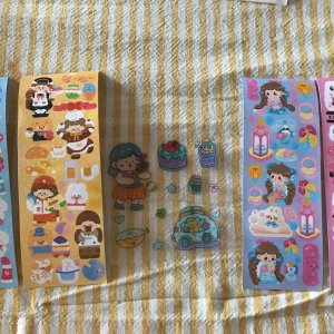 Stickers - Ett gäng färgglada klistermärken med söta figurer och matmotiv. Perfekt för att dekorera anteckningsböcker eller brev. Innehåller olika teman som bakverk, drycker och tecknade karaktärer. Klistermärkena har en lekfull och charmig stil med pastellfärger och glada ansikten.