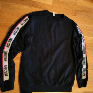 Svart sweatshirt från Moschino - Snygg svart sweatshirt från Moschino med märkets logga längs ärmarna. Tröjan har en klassisk rund hals och ribbade muddar vid ärmslut och nederkant. Perfekt för en stilren och avslappnad look. Nypris ca 3000kr