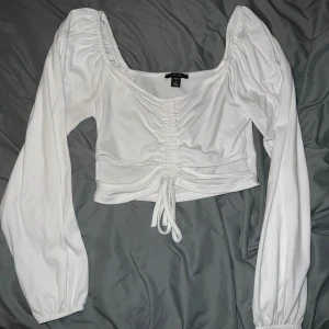 Vit ribbad topp från Amisu - Säljer en snygg vit ribbad topp från Amisu med långa ärmar och knytning framtill. Toppen är croppad och har en smickrande passform med rynkdetaljer vid bysten. Perfekt för en stilren look!