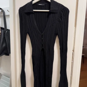 Svart ribbad tröja från Gina Tricot - Snygg svart ribbad tröja från Gina Tricot med v-ringning och knappar framtill. Tröjan har långa ärmar och en klassisk krage. Öppningar i sidor. 