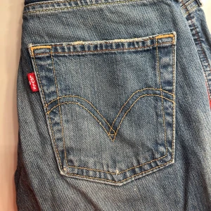 Blå jeans med röda detaljer - Snygga blå jeans från Levis med coola röda detaljer längs sidorna. Klassisk femficksdesign och slitna detaljer för en trendig look. Perfekta för en avslappnad stil.