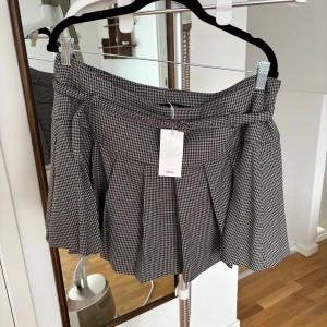 Rutig kortkjol från Mango - Snygg rutig kortkjol från MNG med ett klassiskt mönster i svart och vitt. Kjolen har ett bälte i midjan och veckade detaljer som ger en stilren look. Perfekt för att skapa en chic outfit.