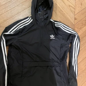 Svart vindjacka från Adidas - Snygg svart vindjacka från Adidas med klassiska vita ränder längs ärmarna och en halv dragkedja framtill. Jackan har en huva och det ikoniska Adidas-loggan på bröstet. Perfekt för sportiga dagar eller en avslappnad stil.
