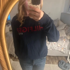 Mörkblå tröja från Tommy Hilfiger - Säljer en snygg mörkblå tröja från Tommy Hilfiger med röd logga på bröstet. Tröjan har långa ärmar och en klassisk passform. Perfekt för en avslappnad stil.