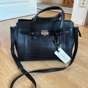 Svart väska från Fiorelli - Svart handväska från märket Fiorelli. Aldrig använd (tag sitter kvar), men små små defekter på handtaget (se sista bilden). Nypris 82,90 EUR ≈ 950 SEK.