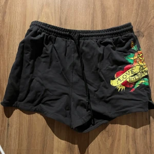 Svarta shorts från Ed Hardy x Urban Outfitters - Säljer ett par svarta shorts från Ed Hardy x Urban Outfitters med färgglatt tryck och text. De har en elastisk midja med dragsko för justerbar passform. Perfekta för en avslappnad stil.