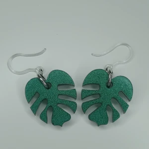 Gröna örhängen i form av monstera-blad - Snygga gröna örhängen formade som monstera-blad. De är mörkgröna glittriga längd ca 3 cm. Öronkroken är gjord I plast 