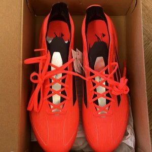 Adidas F50 Elite FG Fotbollsskor - Säljer ett par Adidas F50 Elite FG fotbollsskor i en iögonfallande orange färg med svarta detaljer. Skorna har snörning och är designade för att ge optimal prestanda på fotbollsplanen.