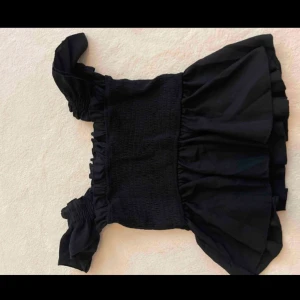 Svart volangtopp - Snygg svart topp med volangdetaljer och smockad överdel. Perfekt för en trendig look med sina breda axelband och volangkant nedtill. 