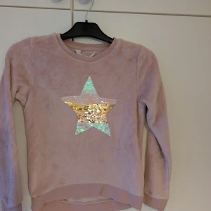 Rosa tröja med paljettstjärna - Säljer en söt rosa tröja med en stor glittrig stjärna i paljetter på framsidan. Tröjan har långa ärmar och är perfekt för att lysa upp vardagen med lite glitter.