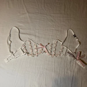  Victoria's Secret bh - Elegant vit bh från Victoria's Secret med vacker blommig spets och en söt rosa rosett framtill. Justerbara axelband för perfekt passform. Perfekt för att känna sig extra fin.