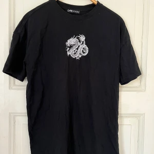 Svart t-shirt med drakmotiv från New yorker  - Snygg svart t-shirt från New yorker med ett coolt vitt drakmotiv på bröstet. Perfekt för en avslappnad stil. Kortärmad och i mjukt material för extra komfort.