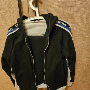 Svart och grå huvtröja/byxod från Diesel - Snygg huvtröja/byxor från Diesel i svart och grått med blå och vit textdetalj på ärmarna. Tröjan har dragkedja och är tillverkad i 100% bomull, vilket gör den bekväm och mjuk. Perfekt för en avslappnad stil.